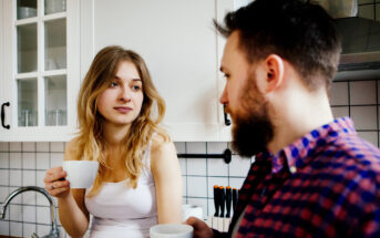 A woman with long blonde hair in a white tank top and a man with a beard in a plaid shirt are standing in a kitchen, holding white mugs. The kitchen has white cabinets and a tiled backsplash. They appear to be engaged in a conversation.