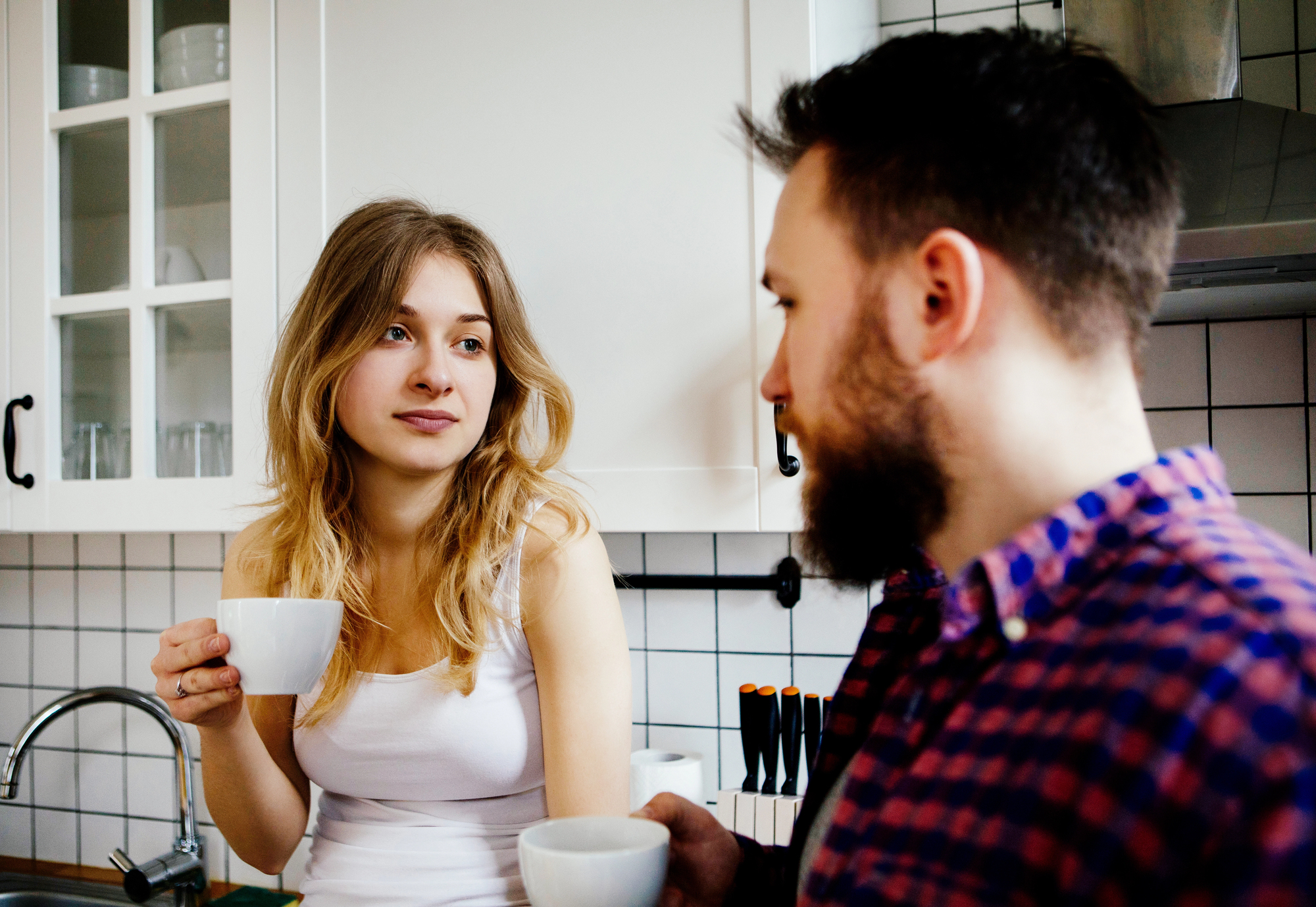 A woman with long blonde hair in a white tank top and a man with a beard in a plaid shirt are standing in a kitchen, holding white mugs. The kitchen has white cabinets and a tiled backsplash. They appear to be engaged in a conversation.