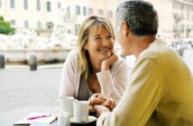 A woman and a man are sitting at an outdoor café, smiling at each other and holding hands. Two cups of coffee are on the table, with a blurred city square visible in the background.