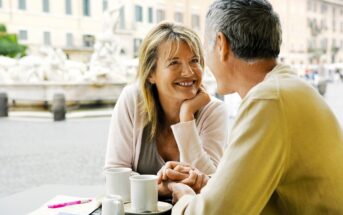 A woman and a man are sitting at an outdoor café, smiling at each other and holding hands. Two cups of coffee are on the table, with a blurred city square visible in the background.