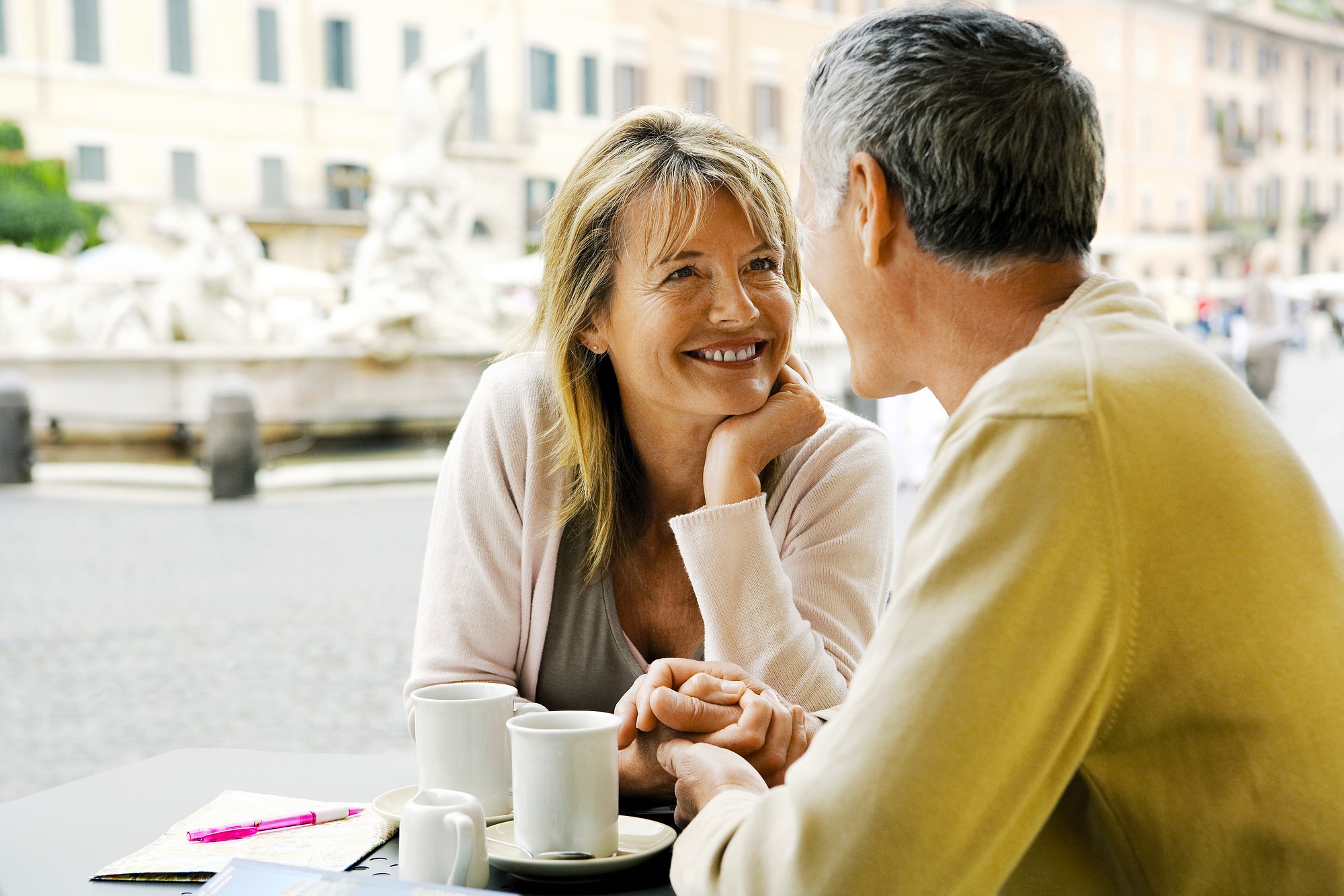 A woman and a man are sitting at an outdoor café, smiling at each other and holding hands. Two cups of coffee are on the table, with a blurred city square visible in the background.