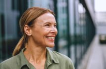 A woman with light brown hair tied back smiles while looking to the side. She is wearing a green blouse and standing outdoors with a blurred modern building in the background.