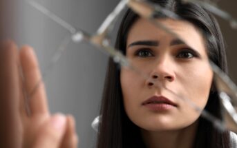 A woman with long dark hair looks intently at a broken mirror, with sharp reflections creating fragmented images of her face. Her expression appears thoughtful and contemplative.