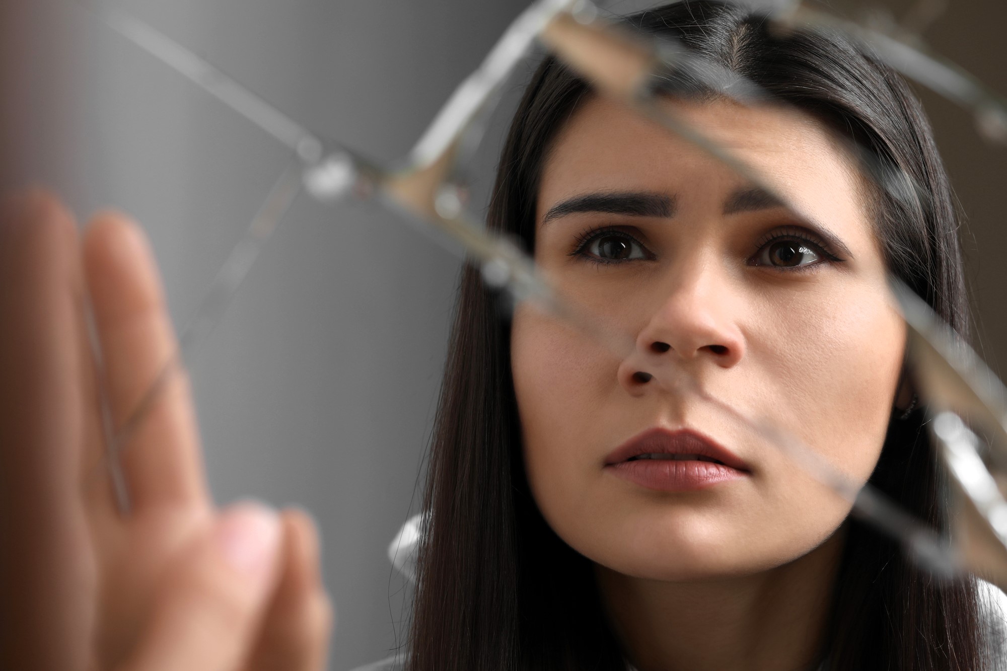 A woman with long dark hair looks intently at a broken mirror, with sharp reflections creating fragmented images of her face. Her expression appears thoughtful and contemplative.