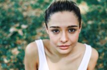 A young woman with dark hair is looking up at the camera. She is outdoors, surrounded by greenery, and wearing a white tank top. The image captures her from the shoulders up, with a blurred background of leaves and grass.