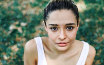 A young woman with dark hair is looking up at the camera. She is outdoors, surrounded by greenery, and wearing a white tank top. The image captures her from the shoulders up, with a blurred background of leaves and grass.