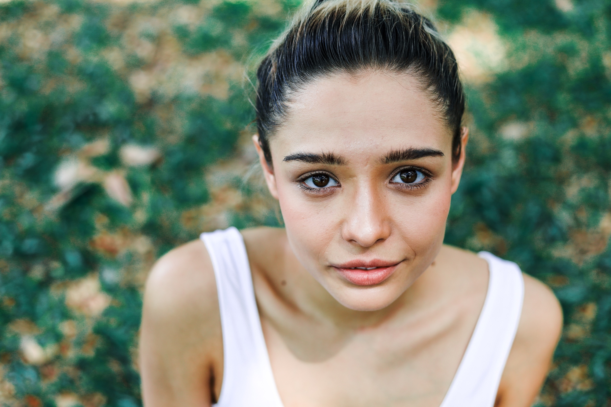 A young woman with dark hair is looking up at the camera. She is outdoors, surrounded by greenery, and wearing a white tank top. The image captures her from the shoulders up, with a blurred background of leaves and grass.