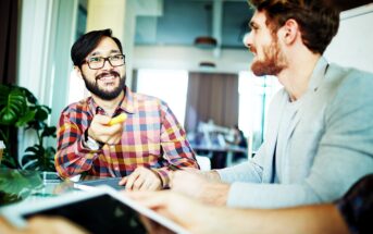 Two men are sitting at a table during a meeting. The man on the left, wearing glasses and a plaid shirt, is smiling and gesturing with a pen. The man on the right, with a beard, is listening attentively. A tablet is visible in the foreground.