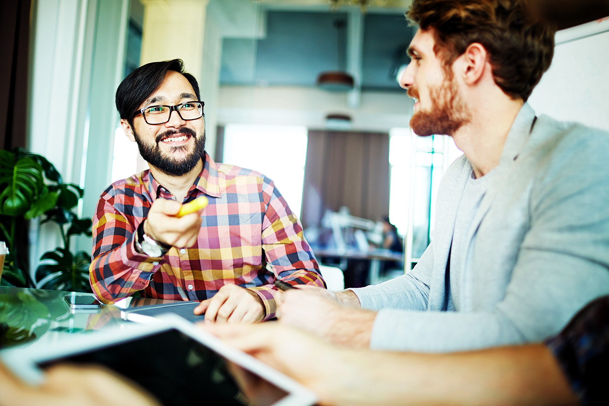 Two men are sitting at a table during a meeting. The man on the left, wearing glasses and a plaid shirt, is smiling and gesturing with a pen. The man on the right, with a beard, is listening attentively. A tablet is visible in the foreground.