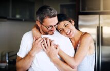 A woman is embracing a man from behind in a kitchen, both smiling warmly. The man wears glasses and a white shirt, while the woman is in a sleeveless top. They appear happy and relaxed, with a modern kitchen setting in the background.