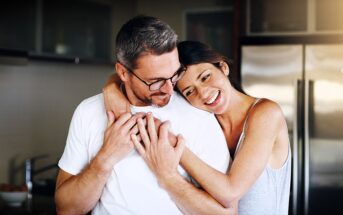 A woman is embracing a man from behind in a kitchen, both smiling warmly. The man wears glasses and a white shirt, while the woman is in a sleeveless top. They appear happy and relaxed, with a modern kitchen setting in the background.