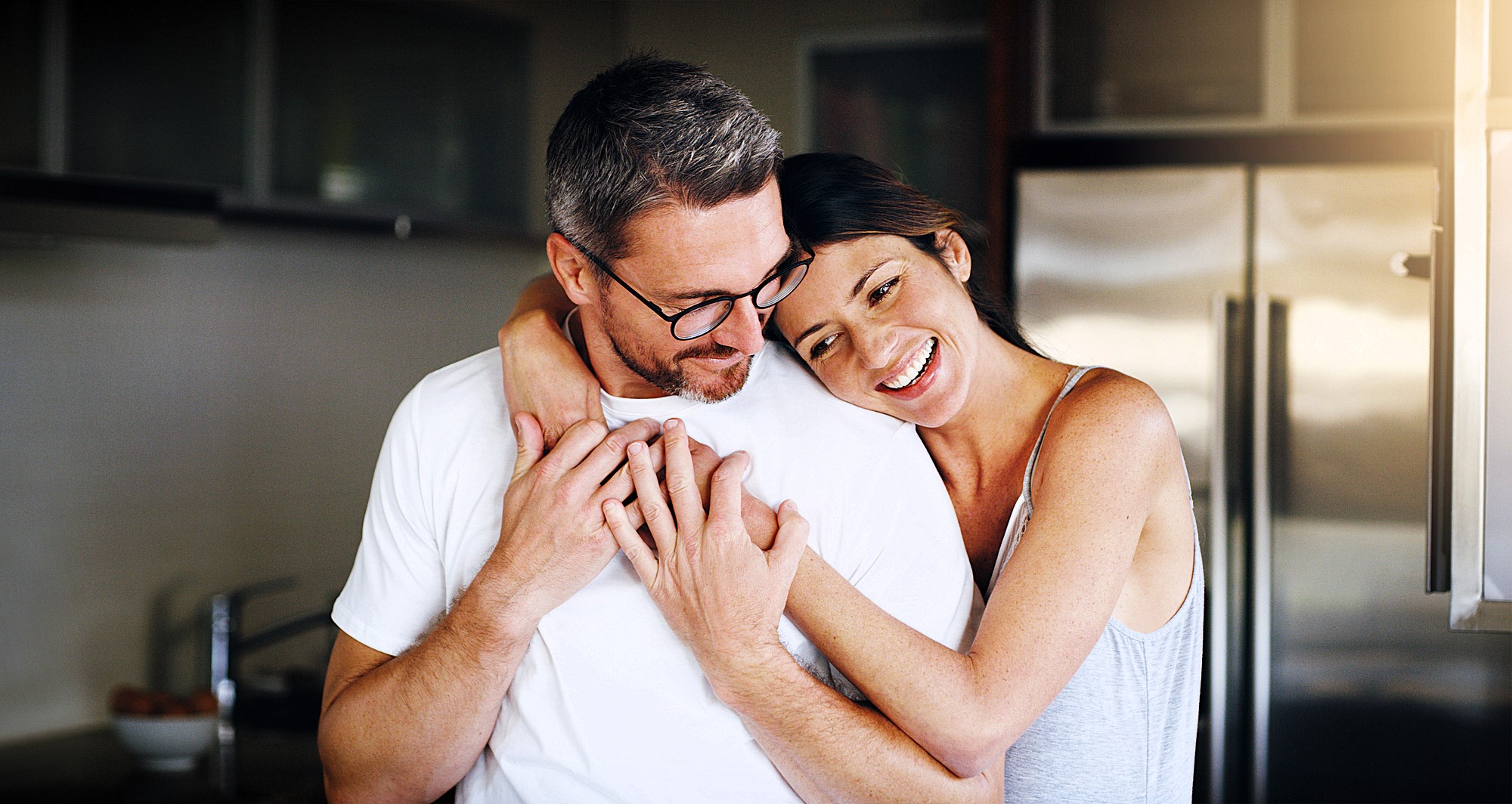 A woman is embracing a man from behind in a kitchen, both smiling warmly. The man wears glasses and a white shirt, while the woman is in a sleeveless top. They appear happy and relaxed, with a modern kitchen setting in the background.