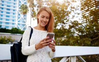 A woman in a white sweater stands outdoors, smiling while looking at her phone. She carries a black bag over her shoulder. The background features a modern building and trees under a clear sky.