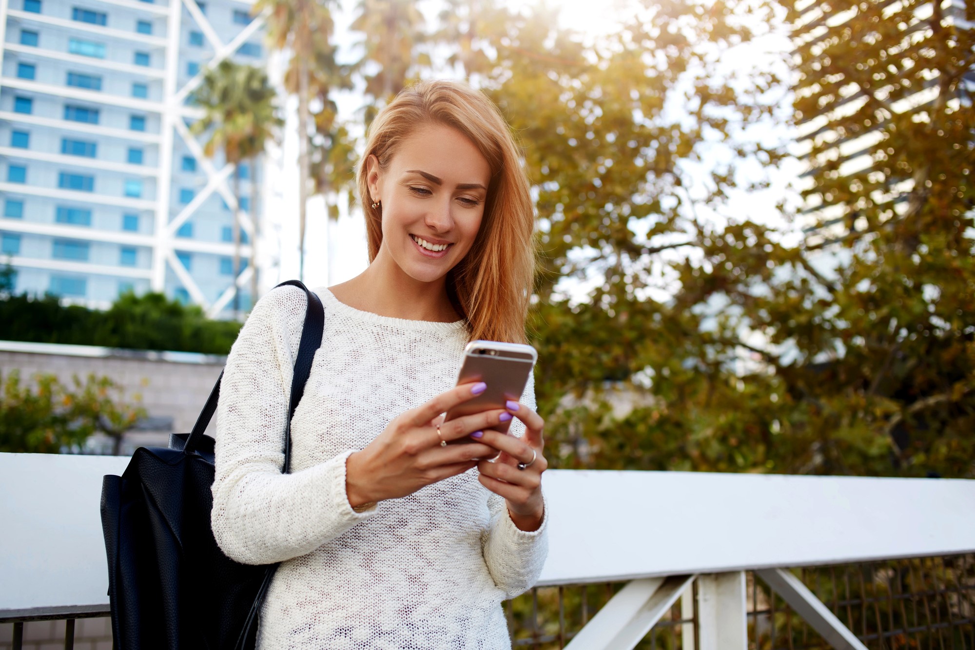 A woman in a white sweater stands outdoors, smiling while looking at her phone. She carries a black bag over her shoulder. The background features a modern building and trees under a clear sky.