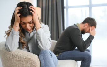 A woman sits on a chair with her head in her hands, visibly upset. In the background, a man sits on a couch, also with his head in his hands, appearing distressed. The scene suggests a tense or emotional moment between them.