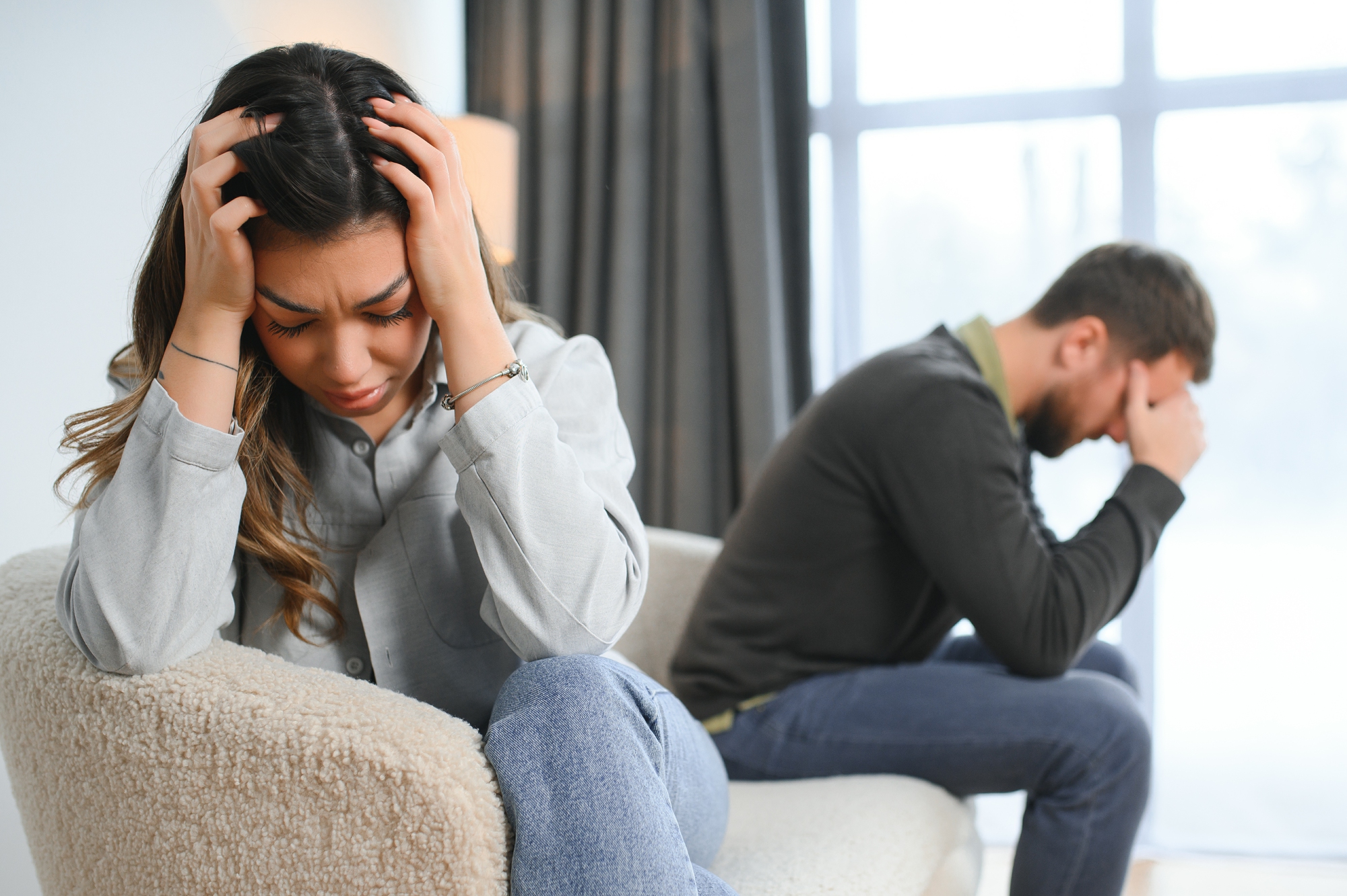 A woman sits on a chair with her head in her hands, visibly upset. In the background, a man sits on a couch, also with his head in his hands, appearing distressed. The scene suggests a tense or emotional moment between them.