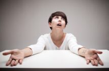 A person with short dark hair sits at a table with eyes looking upward and arms outstretched. They wear a white long-sleeve shirt and appear exasperated or frustrated, against a plain gray background.