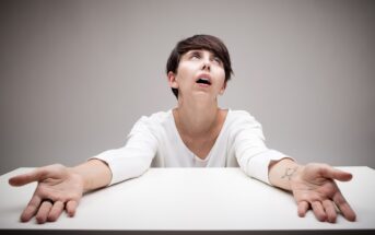 A person with short dark hair sits at a table with eyes looking upward and arms outstretched. They wear a white long-sleeve shirt and appear exasperated or frustrated, against a plain gray background.