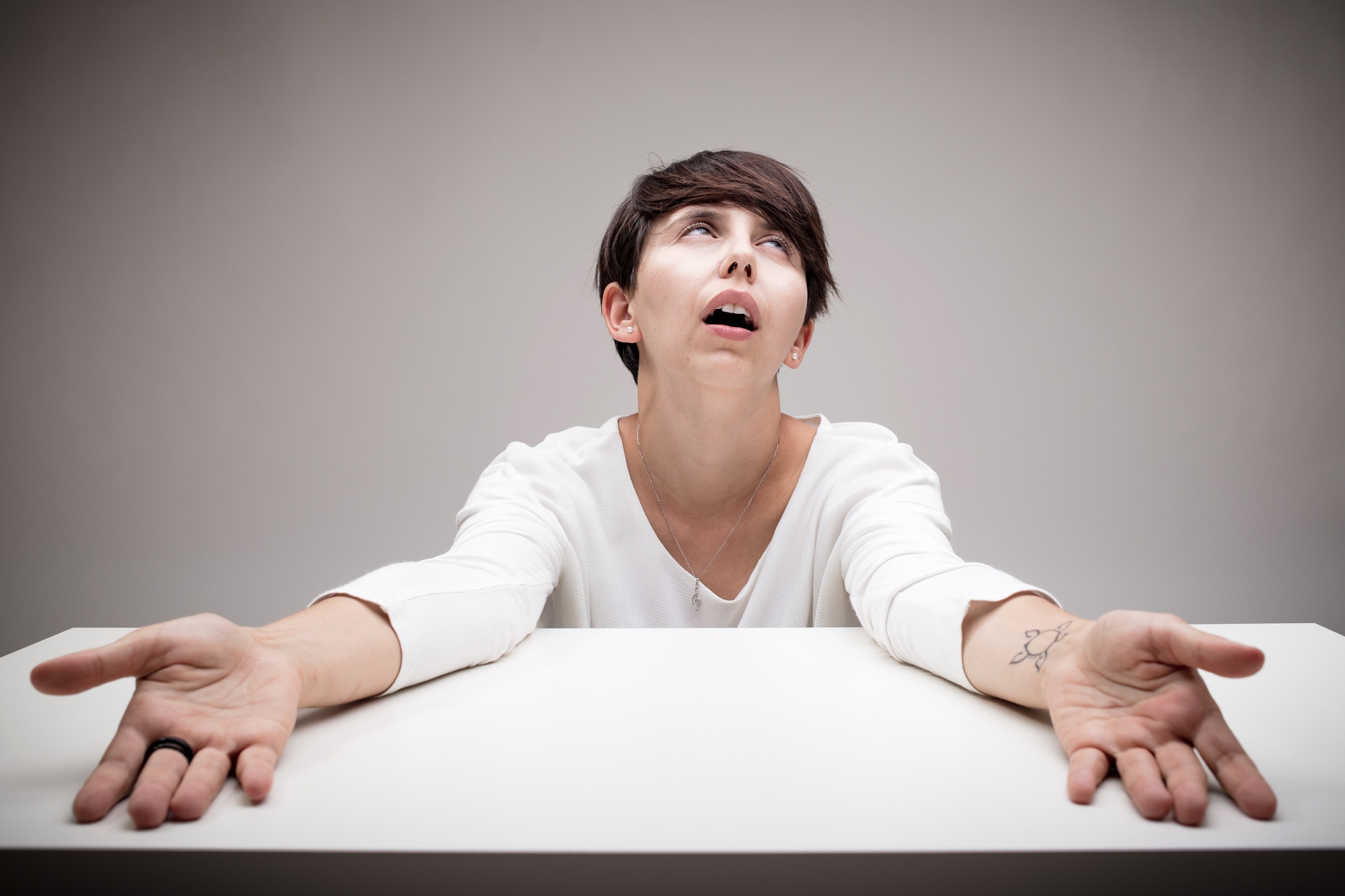 A person with short dark hair sits at a table with eyes looking upward and arms outstretched. They wear a white long-sleeve shirt and appear exasperated or frustrated, against a plain gray background.