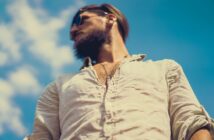 A man with a beard and sunglasses is looking to the side against a clear blue sky. He is wearing a light, buttoned shirt. The perspective is from below, giving a slightly upward angle.