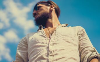 A man with a beard and sunglasses is looking to the side against a clear blue sky. He is wearing a light, buttoned shirt. The perspective is from below, giving a slightly upward angle.