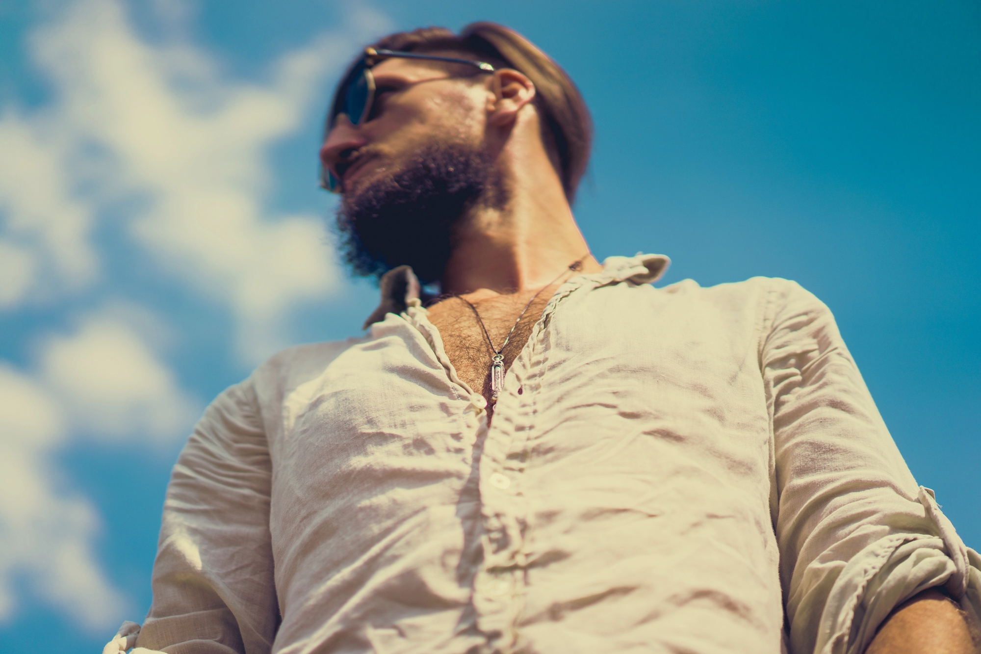 A man with a beard and sunglasses is looking to the side against a clear blue sky. He is wearing a light, buttoned shirt. The perspective is from below, giving a slightly upward angle.