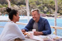 A man and woman enjoy a meal at an outdoor table by the sea. They are smiling at each other, with food and drinks in front of them. The background shows a beach with trees and lounge chairs under umbrellas.