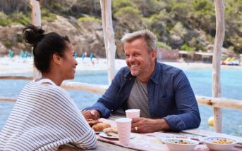 A man and woman enjoy a meal at an outdoor table by the sea. They are smiling at each other, with food and drinks in front of them. The background shows a beach with trees and lounge chairs under umbrellas.