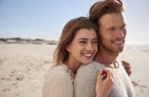 A couple stands on a sandy beach, smiling and embracing each other. The woman is dressed in a light sweater, and the man in a gray one. The sky is clear, and the sea is visible in the background, creating a serene and joyful atmosphere.