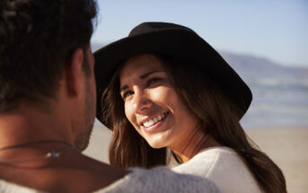 A woman wearing a black hat smiles warmly at a man on a beach. The sun is shining, and the ocean and mountains are visible in the background. They appear to be in a relaxed and happy moment.