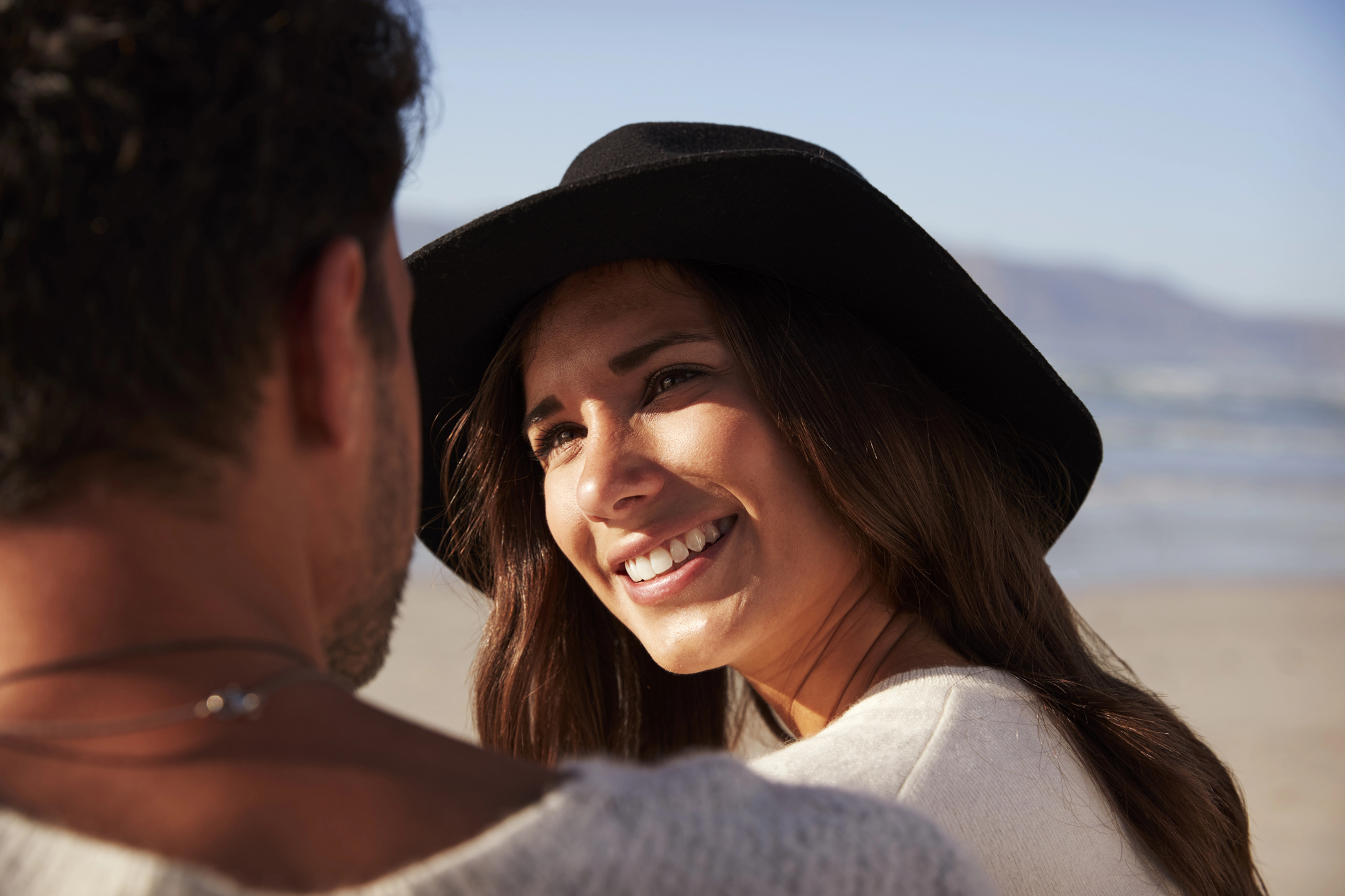 A woman wearing a black hat smiles warmly at a man on a beach. The sun is shining, and the ocean and mountains are visible in the background. They appear to be in a relaxed and happy moment.