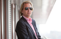 A person with blonde hair wearing sunglasses and a pinstripe suit with a pink shirt stands outside near a building, looking into the distance.