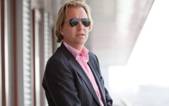 A person with blonde hair wearing sunglasses and a pinstripe suit with a pink shirt stands outside near a building, looking into the distance.