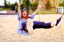 A person with long red hair and a hat swings joyfully on a playground swing, wearing a blue shirt and black pants. The background features a sandy ground and blurred greenery on a sunny day.