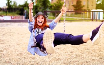 A person with long red hair and a hat swings joyfully on a playground swing, wearing a blue shirt and black pants. The background features a sandy ground and blurred greenery on a sunny day.