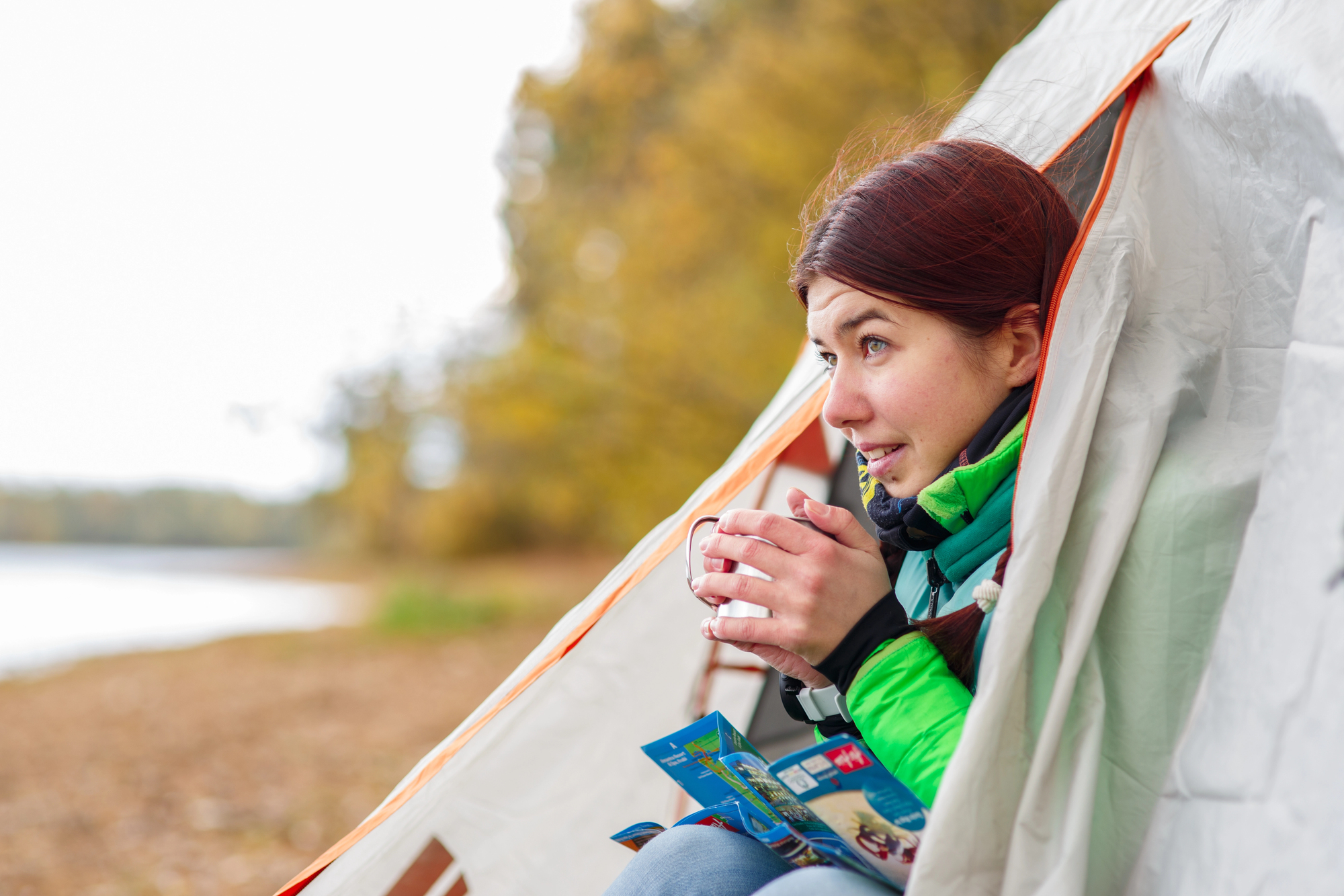 A woman sits at the entrance of a tent by a lake, holding a mug. She wears a green jacket and looks content, with autumn trees in the background. A map rests on her lap.