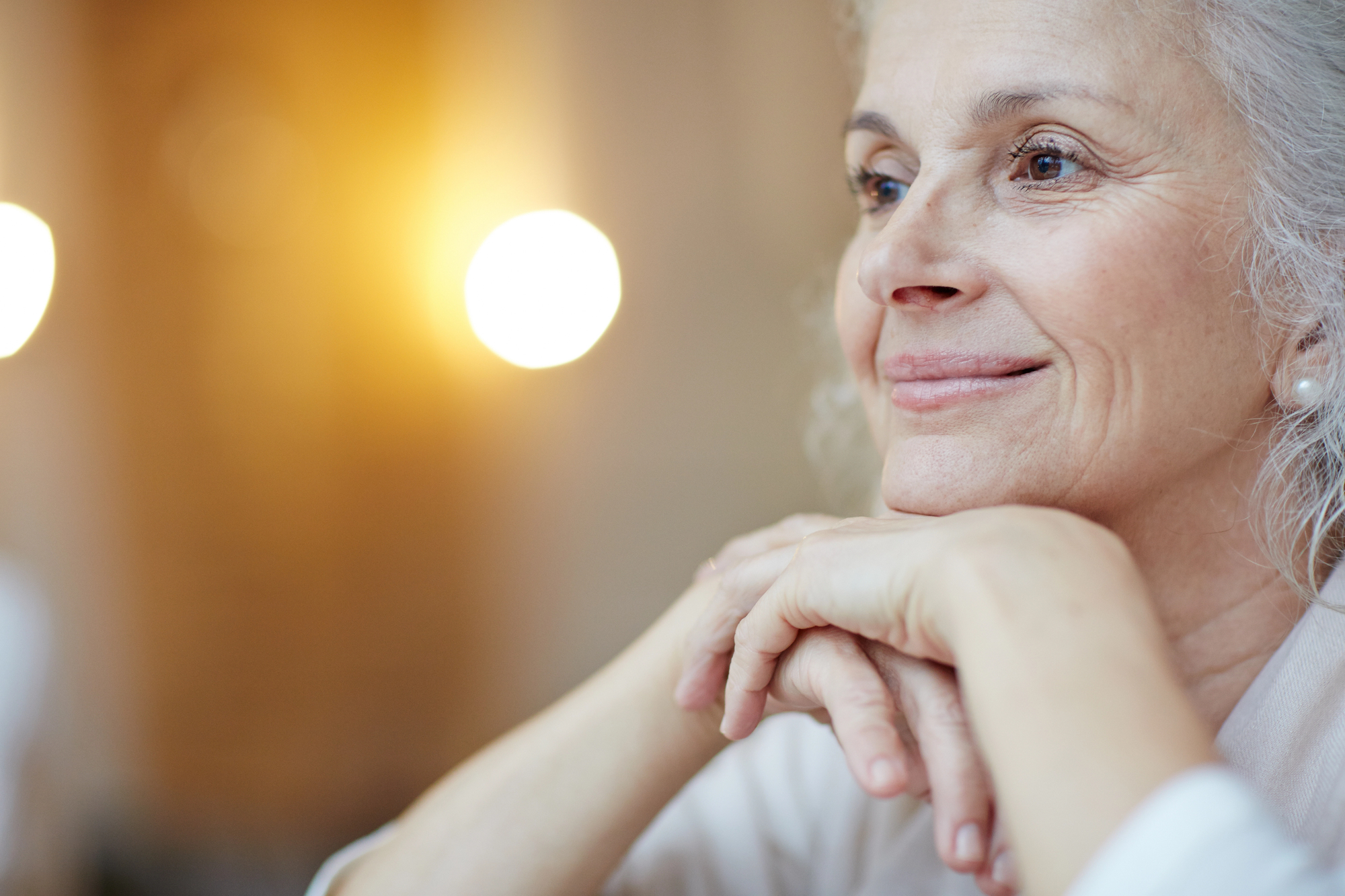 An elderly woman with gray hair and a gentle smile rests her chin on her hands, looking thoughtfully to the side. The background is softly blurred with warm lighting.