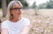 A middle-aged woman with short blonde hair and glasses is sitting outdoors in a park, wearing a white shirt. She looks thoughtfully into the distance, with blurred trees and grass in the background.