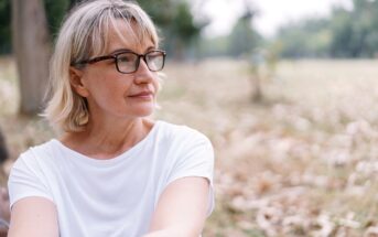 A middle-aged woman with short blonde hair and glasses is sitting outdoors in a park, wearing a white shirt. She looks thoughtfully into the distance, with blurred trees and grass in the background.