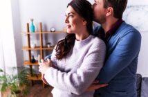 A couple stands in a cozy room, with the man gently hugging the woman from behind and kissing her head. She smiles softly while looking to the side. Shelving with decorative items and a plant are visible in the background.