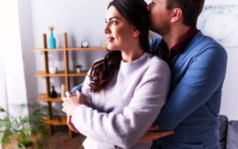 A couple stands in a cozy room, with the man gently hugging the woman from behind and kissing her head. She smiles softly while looking to the side. Shelving with decorative items and a plant are visible in the background.