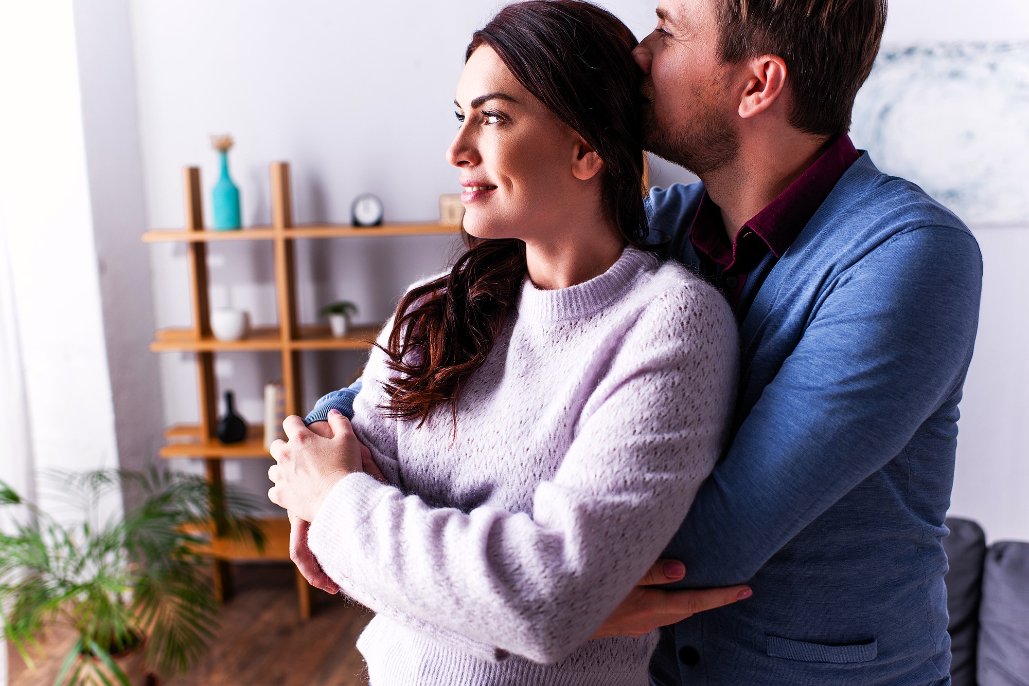 A couple stands in a cozy room, with the man gently hugging the woman from behind and kissing her head. She smiles softly while looking to the side. Shelving with decorative items and a plant are visible in the background.