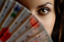 Close-up of a person partially hiding their face with a colorful fan. Only one eye and part of the forehead are visible. The fan has floral patterns with red, green, and blue hues. The focus is on the eye, which has striking details.