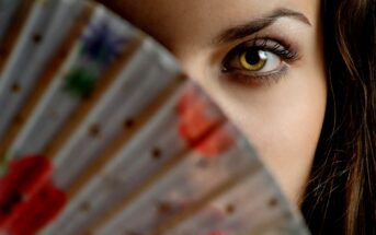 Close-up of a person partially hiding their face with a colorful fan. Only one eye and part of the forehead are visible. The fan has floral patterns with red, green, and blue hues. The focus is on the eye, which has striking details.