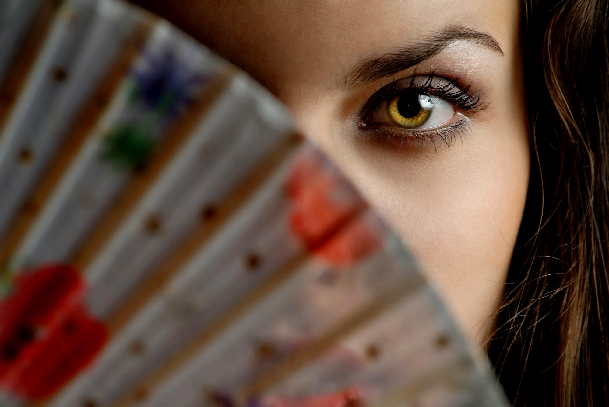 Close-up of a person partially hiding their face with a colorful fan. Only one eye and part of the forehead are visible. The fan has floral patterns with red, green, and blue hues. The focus is on the eye, which has striking details.