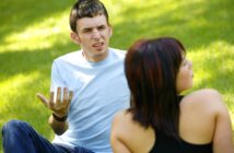 A man in a light blue shirt, sitting on grass, gestures with an exasperated expression. A woman with short dark hair, wearing a black top, sits nearby with her back to the camera. The setting is a sunny outdoor park.