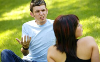 A man in a light blue shirt, sitting on grass, gestures with an exasperated expression. A woman with short dark hair, wearing a black top, sits nearby with her back to the camera. The setting is a sunny outdoor park.