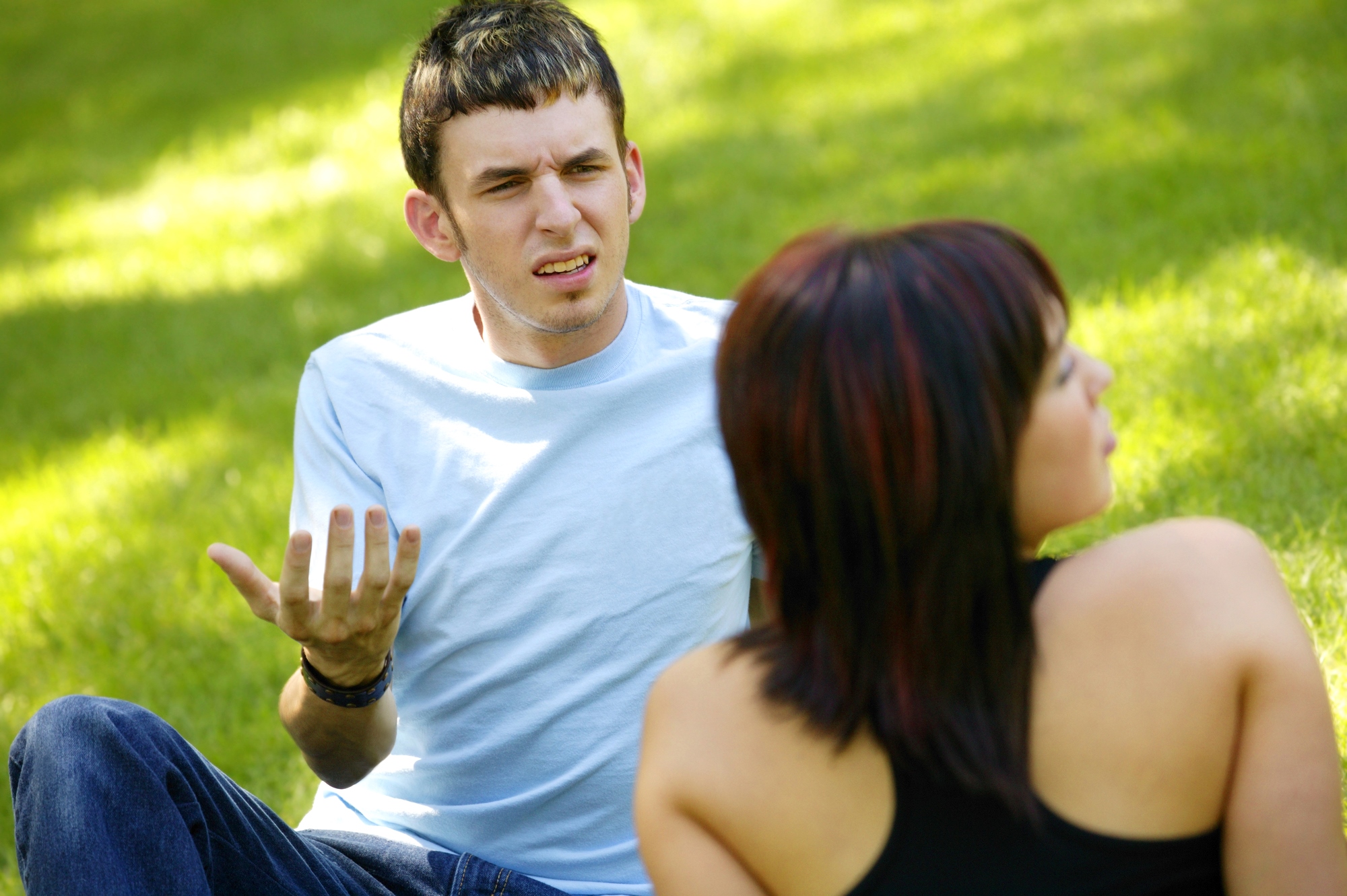 A man in a light blue shirt, sitting on grass, gestures with an exasperated expression. A woman with short dark hair, wearing a black top, sits nearby with her back to the camera. The setting is a sunny outdoor park.