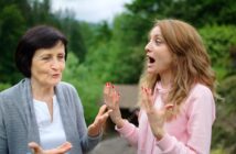 Two women having an animated conversation in an outdoor setting. The older woman, wearing a gray cardigan, gestures with her hands, while the younger woman in a pink hoodie appears surprised, with hands raised. Lush greenery is in the background.