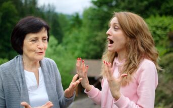 Two women having an animated conversation in an outdoor setting. The older woman, wearing a gray cardigan, gestures with her hands, while the younger woman in a pink hoodie appears surprised, with hands raised. Lush greenery is in the background.